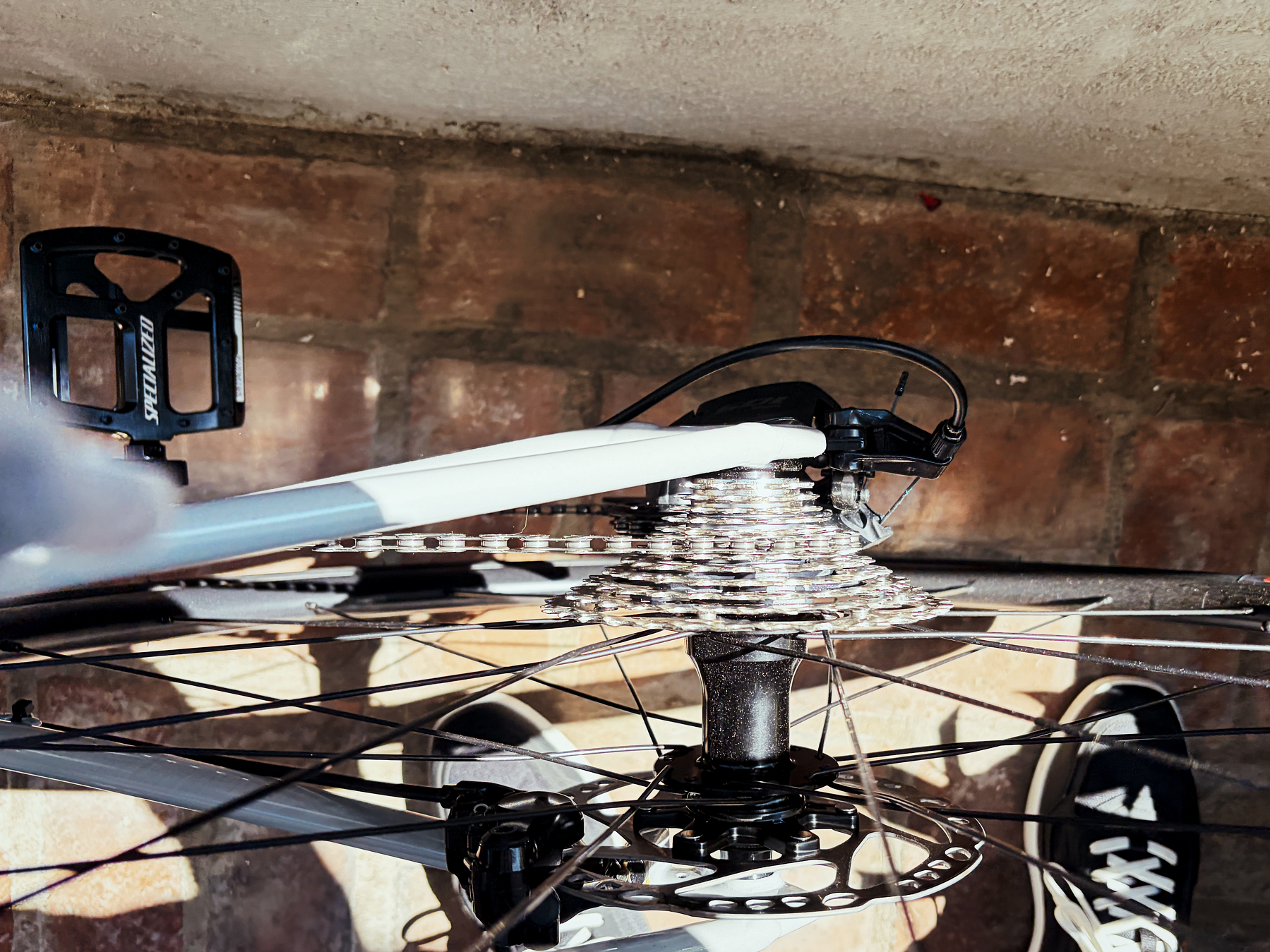 A close-up view of a bicycle's rear cassette, chain, and wheel hub taken from above, with a tiled floor and a person's feet in black sneakers visible in the background.