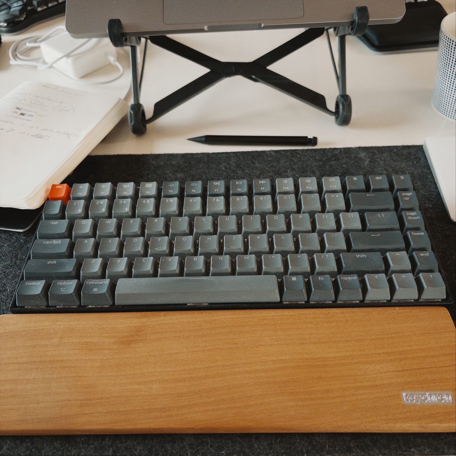 A mechanical keyboard with dark keycaps and an orange escape key sits on a black mat beside a wooden wrist rest, with a German-made black mechanical pencil above it and a laptop on a stand in the background.